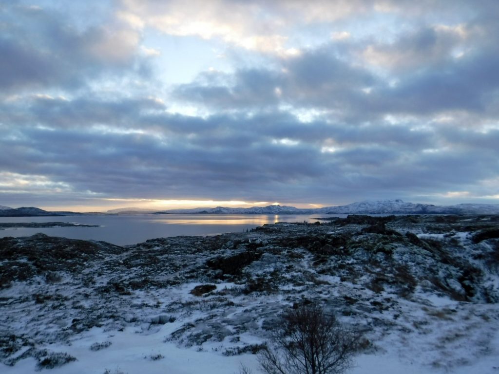 A moody snowy landscape. The golden sun reflects off still water in the distance.