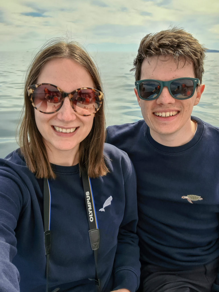 A couple sit smiling on a boat. Their navy blue jumpers have small marine animals knitted on them.