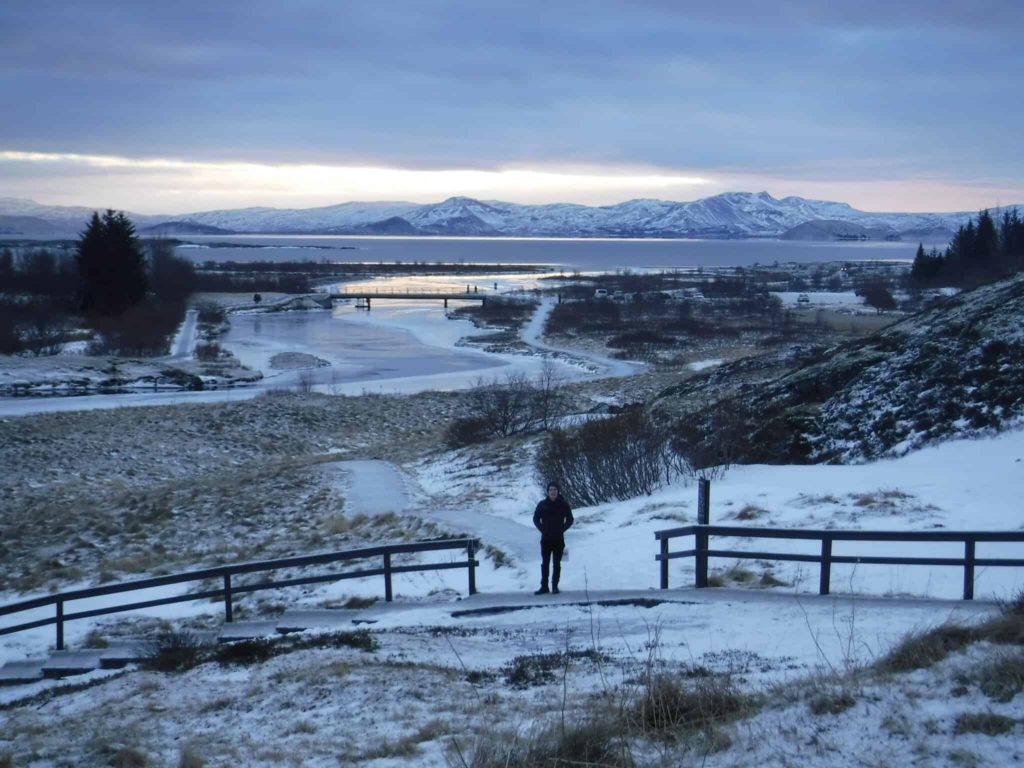 Þingvellir National Park - a boy stands in front of a sweeping landscape. A small bridge crosses an icy stream leading to a calm lake.
