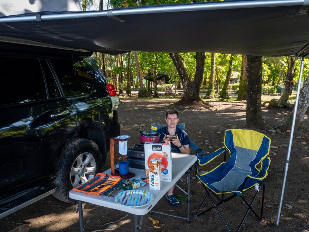 A man sits in a camping chair and enjoys his breakfast. In front of him is a camping stove some breakfast items. He is sitting under an awning by the beach.