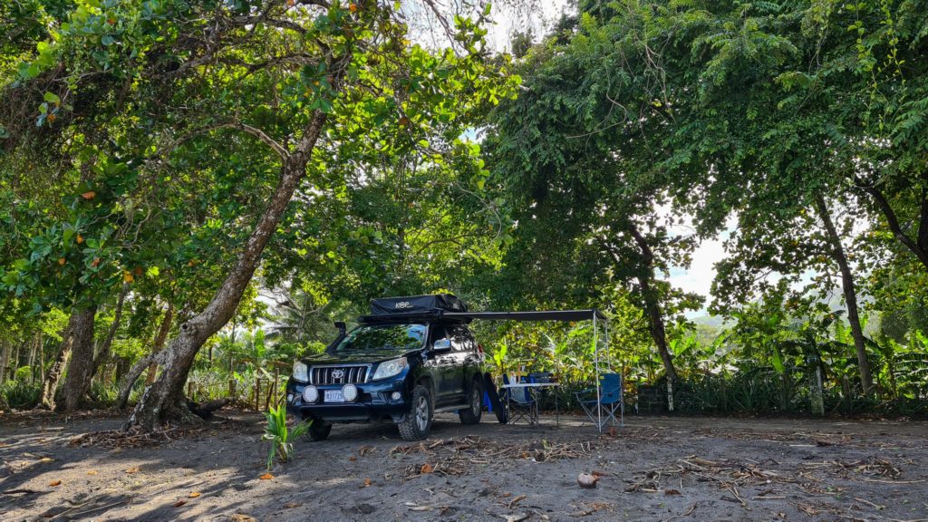 A camper rental car parked by the beach with awning set up. Greenery surrounds the scene.