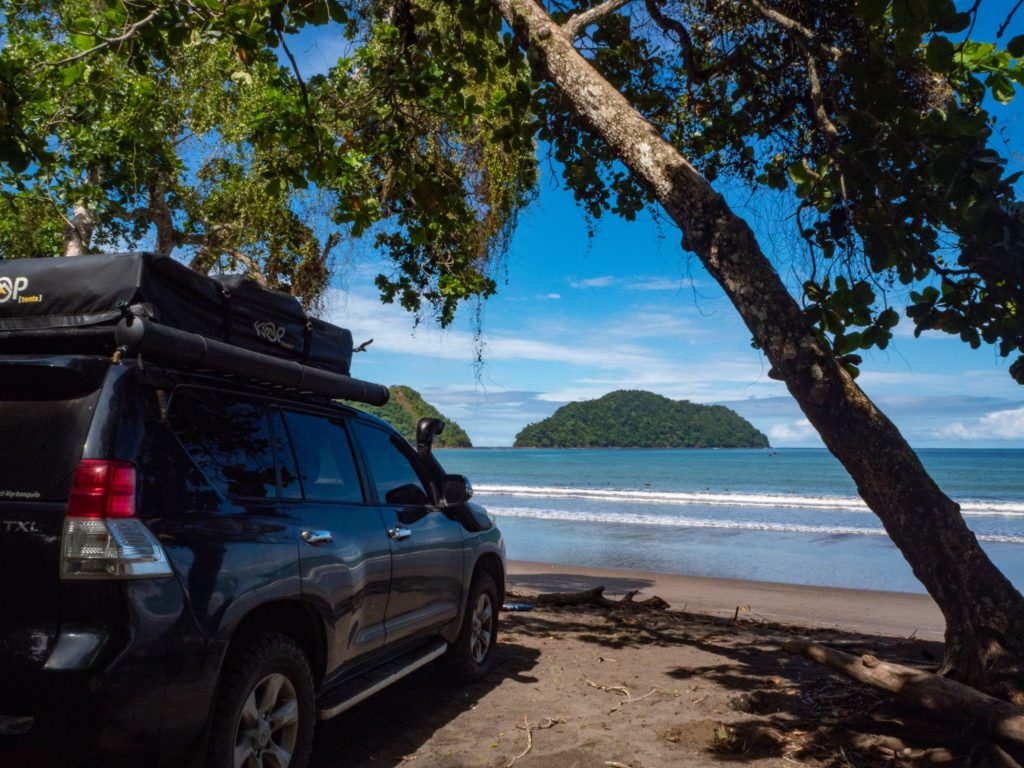 The view of the beach from beside our car. A tree hangs over the car and an island can be seen in the distance.