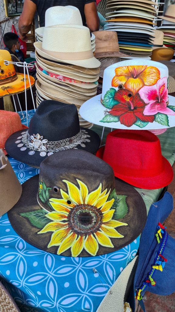 We see a selection of colourful hats on offer at a market in La Paz, Mexico. A couple of these hats have been painted with detailed flowers.