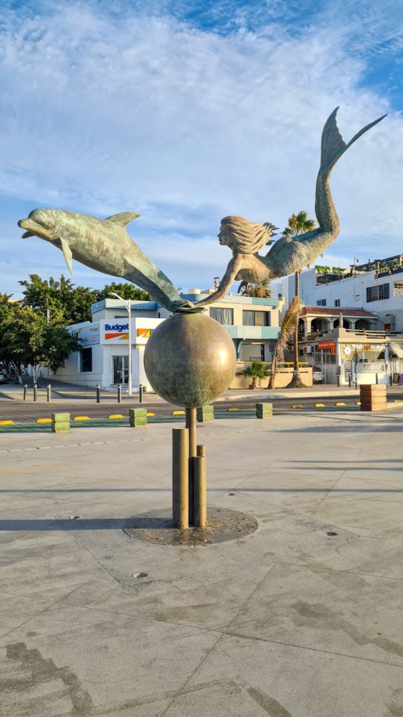 A large metal sculpture stands at the end of a street in La Paz. It depicts a mermaid swimming with a dolphin.