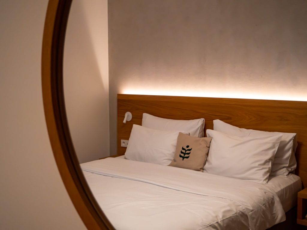 A sleek white bed in the room of an eco-hotel is reflected in a circular wooden mirror. The lighting is warm and a pillow displays a green leaf.