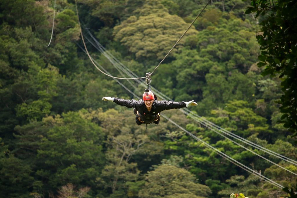 A boy, lying on his front with his arms out either side, ziplines over thick rainforest.