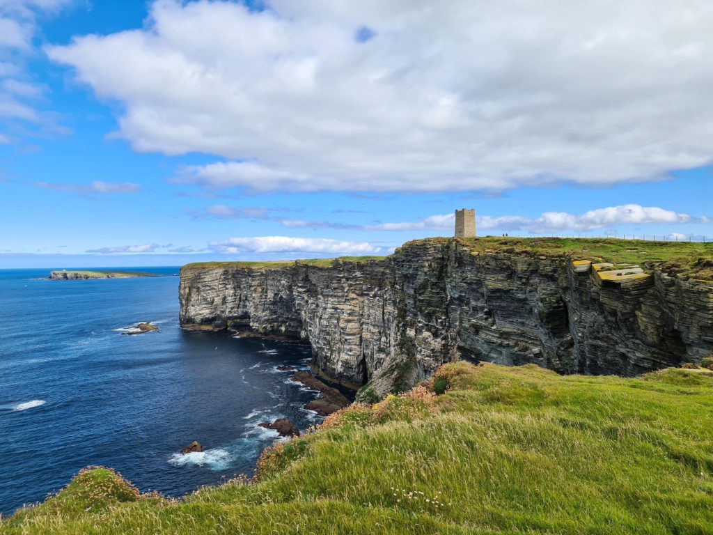 A small tower stands out against the green backdrop of sheer cliffs. The sea stretches out to the horizon.