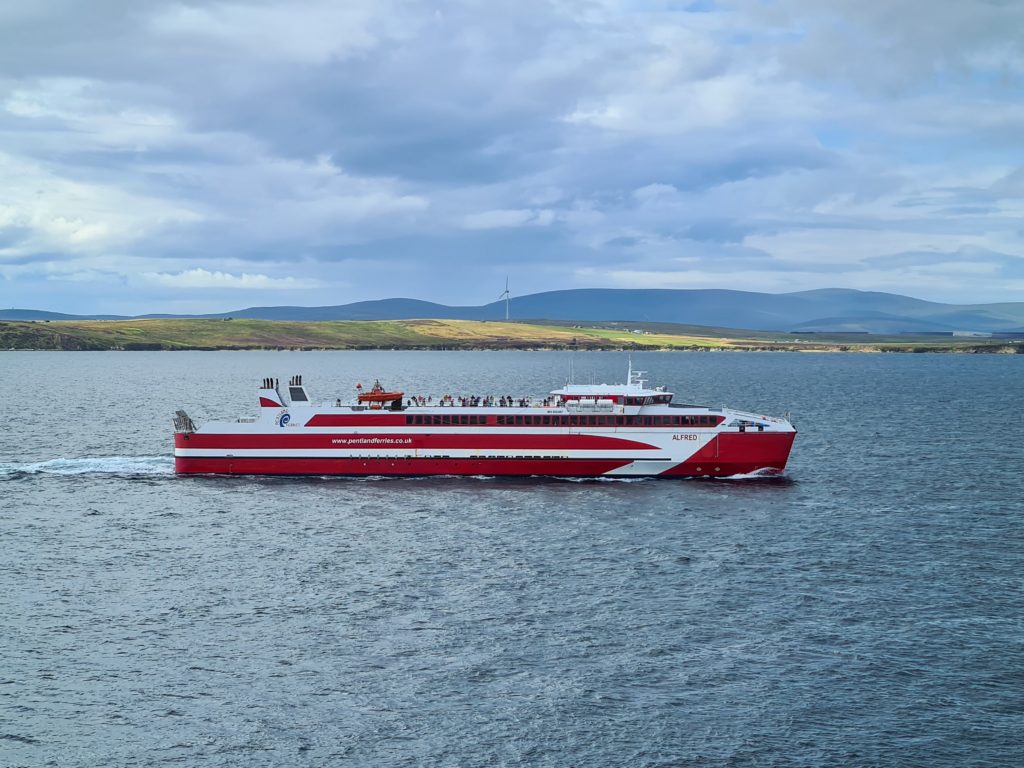 A red ferry baring the name "ALFRED" cruises through the sea. In the background we see green hills and a wind turbine.