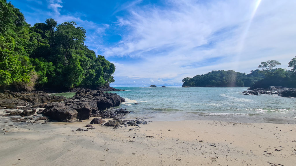 Standing on the sand of Playa Manuel Antonio, we see the light blue waters stretch out to the horizon. Dark rocks and dense forest surround the beach.