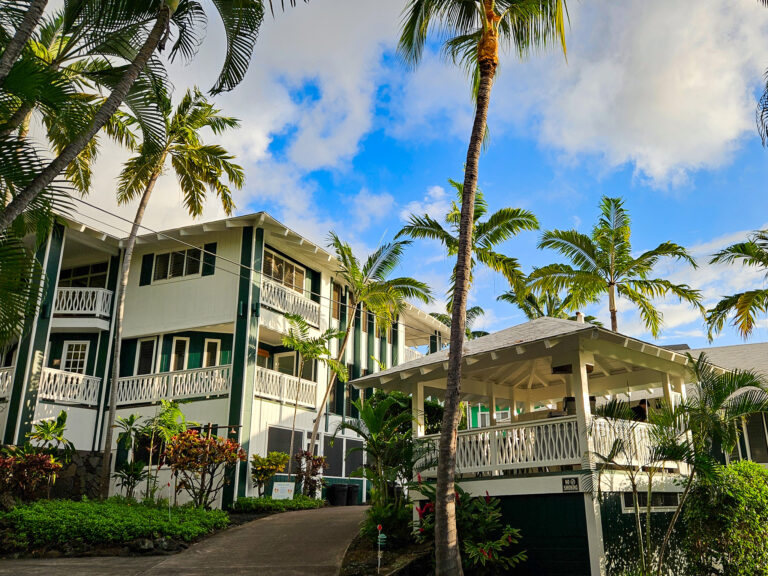 Surrounded by palm trees, Big Island Retreat stands in the sunshine. The buildings are open-air style with balconies and lots of windows.
