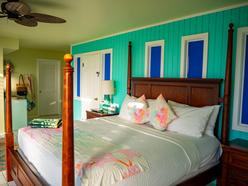 A four poster bed stands in a a hotel room with bright teal walls. The wooden posts cast shadows on the bed.