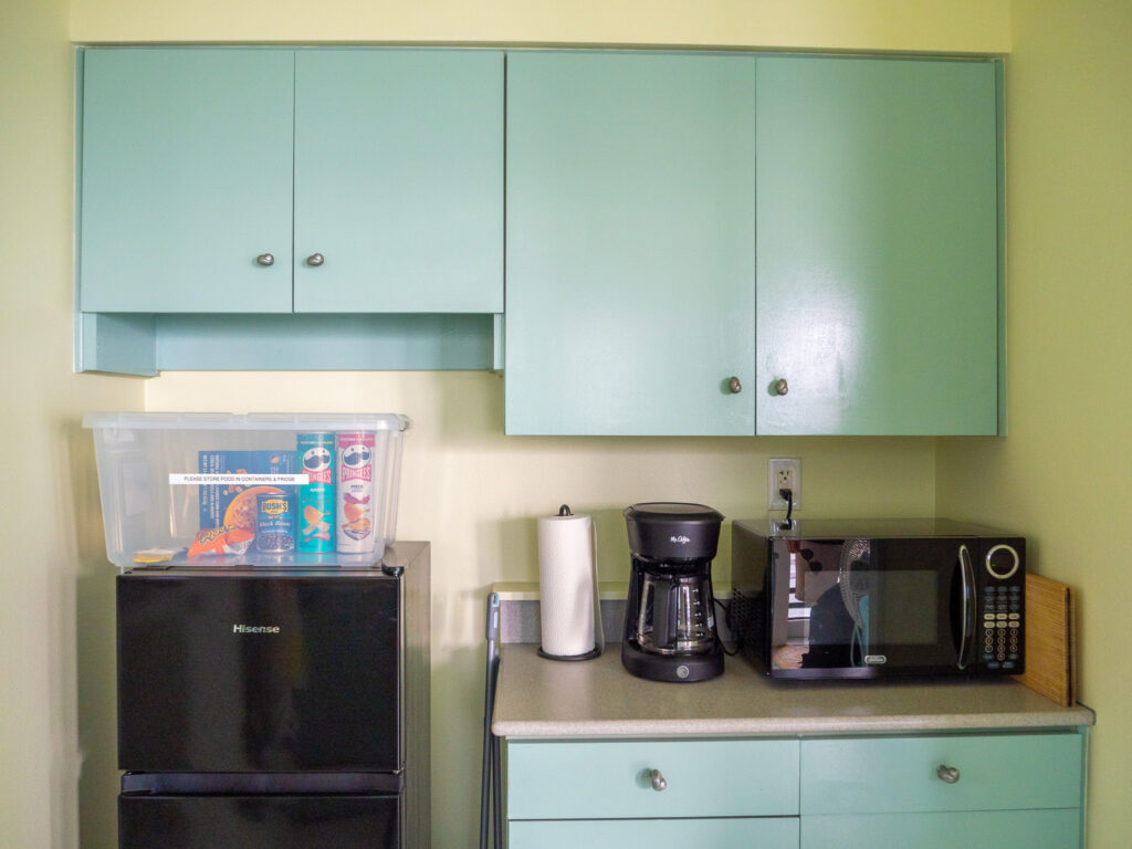 We see a large tub of assorted snacks resting on top of a black fridge-freezer. There are several cupboards close by as well as a coffee maker and microwave.