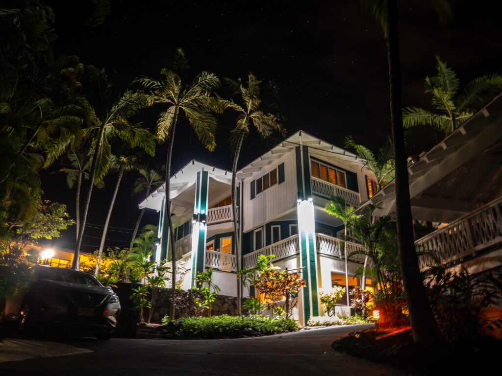 Night time at Big Island Retreat. The white and green building is light up with spot lights and the whole structure is surrounded by palm trees.