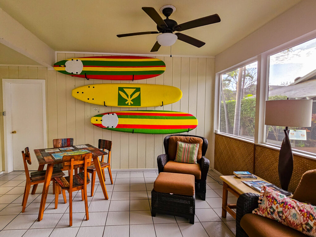 A small communal area has a patchwork wooden table, a comfy armchair, and three large painted surfboards mounted on the back wall.