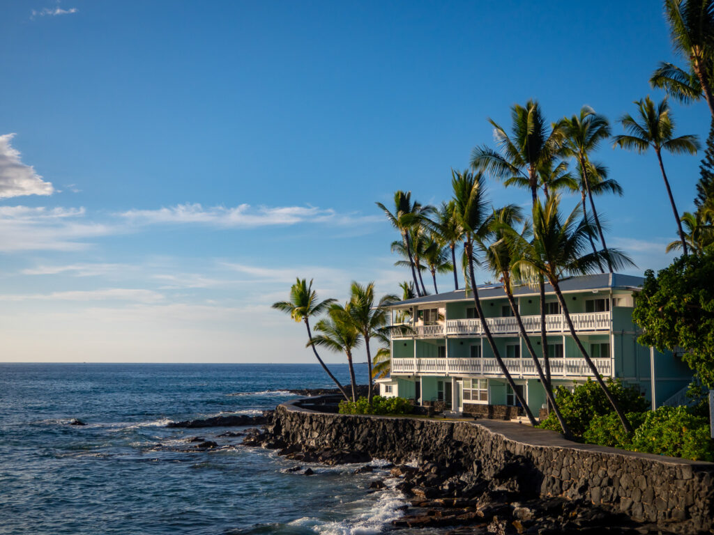 Palm trees surround the charismatic exterior of Kona Tiki Hotel. The sun is setting and the ocean is calm. Each room has a generous balcony and view of the sea.