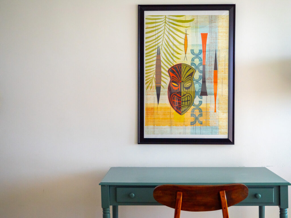 A colourful piece of Hawaiian art hangs from the wall. Below it is a painted wooden desk and chair.