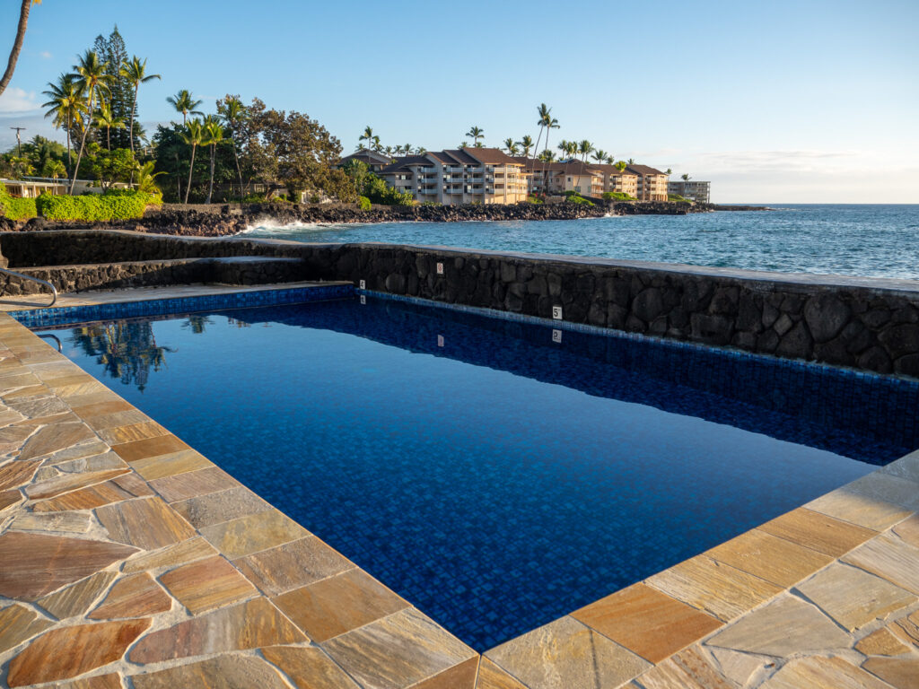 The large pool at Kona Tiki Hotel boasts luxury views of the sea and of the surrounding palm trees. The bottom of the pool is decorated with patterned blue tiles.