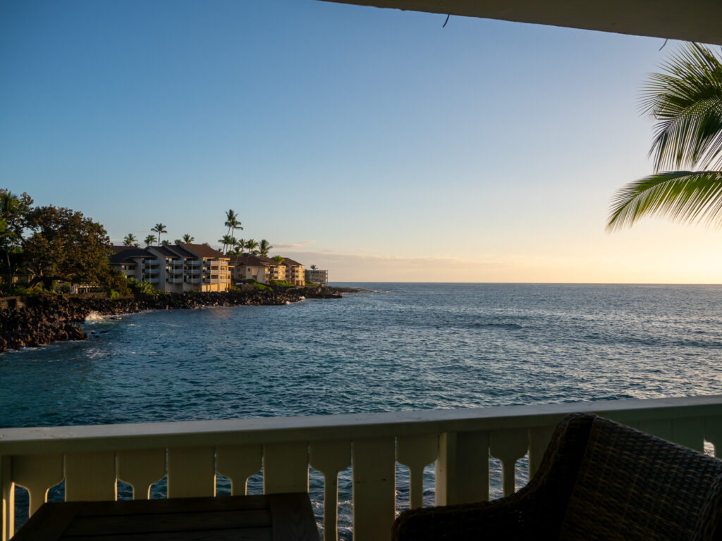 The view of the ocean at sunset from one of the private lanais in Kona Tiki Hotel. A palm tree pokes into shot from the right hand side and the sky is changing from blue to orange.
