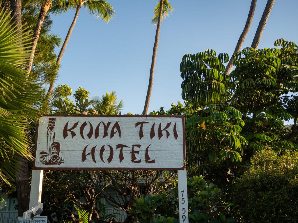 A white sign reads "KONA TIKI HOTEL". It is decorated with hawaiian images and sits against a backdrop of palm trees.