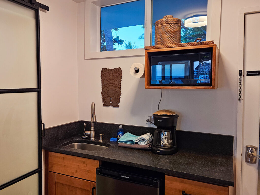 We see a kitchenette complete with sink, coffee maker, microwave and fridge. A wooden carving hangs on the the wall.