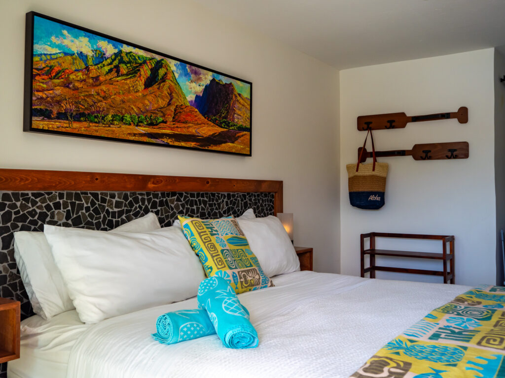 A king sized bed fills the view. The towels and blankets are decorated with animal and pineapple shapes. In the background, a large beach bag hangs on the wall.