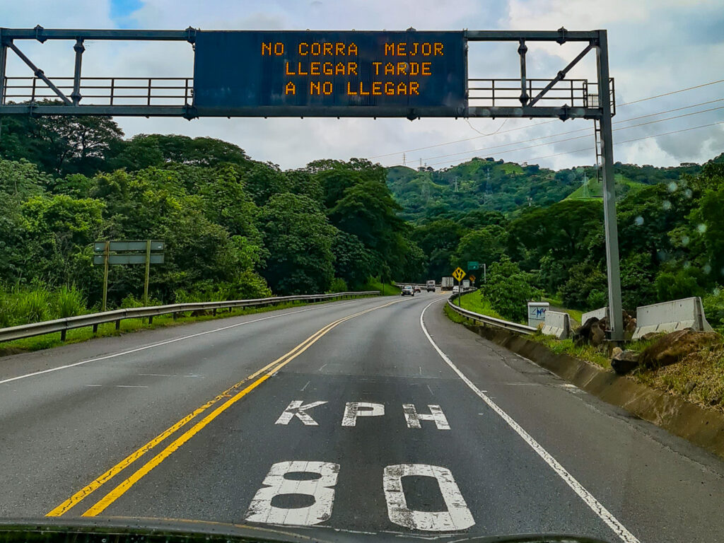 Driving down a road in Costa Rica. Either side of the road is full of greenery. An overhanging road sign reads "No corra, mejor llagar tarde a no llegar", translating to "Don't rush, it's better to be late than not to arrive."