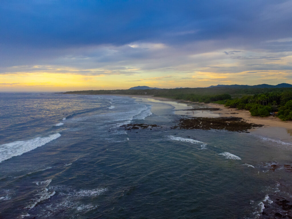 Aerial view across the ocean towards the sunset on a tropical beach. There is a golden light in the sky and a few people paddling in the waves.