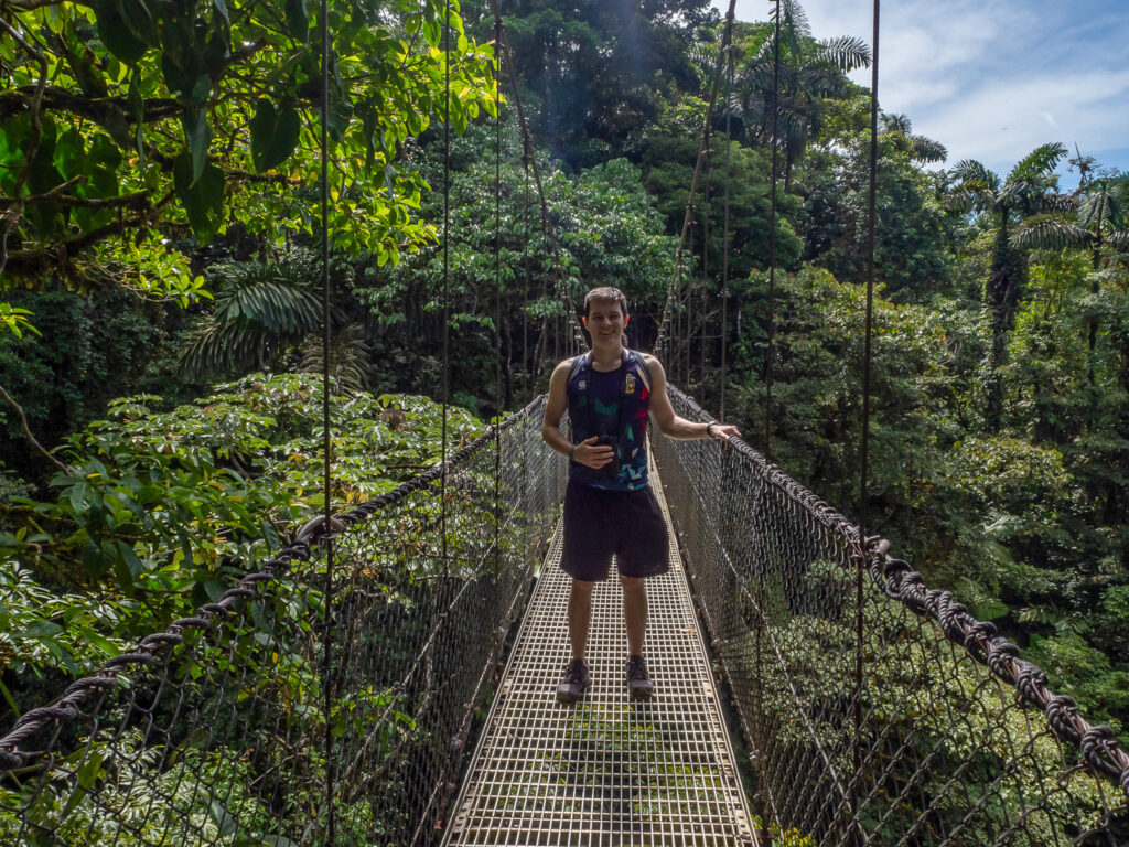 Dan stands on a long hanging bridge surrounded by greenery. The leaves on the trees in the scene are many different shapes and sizes.