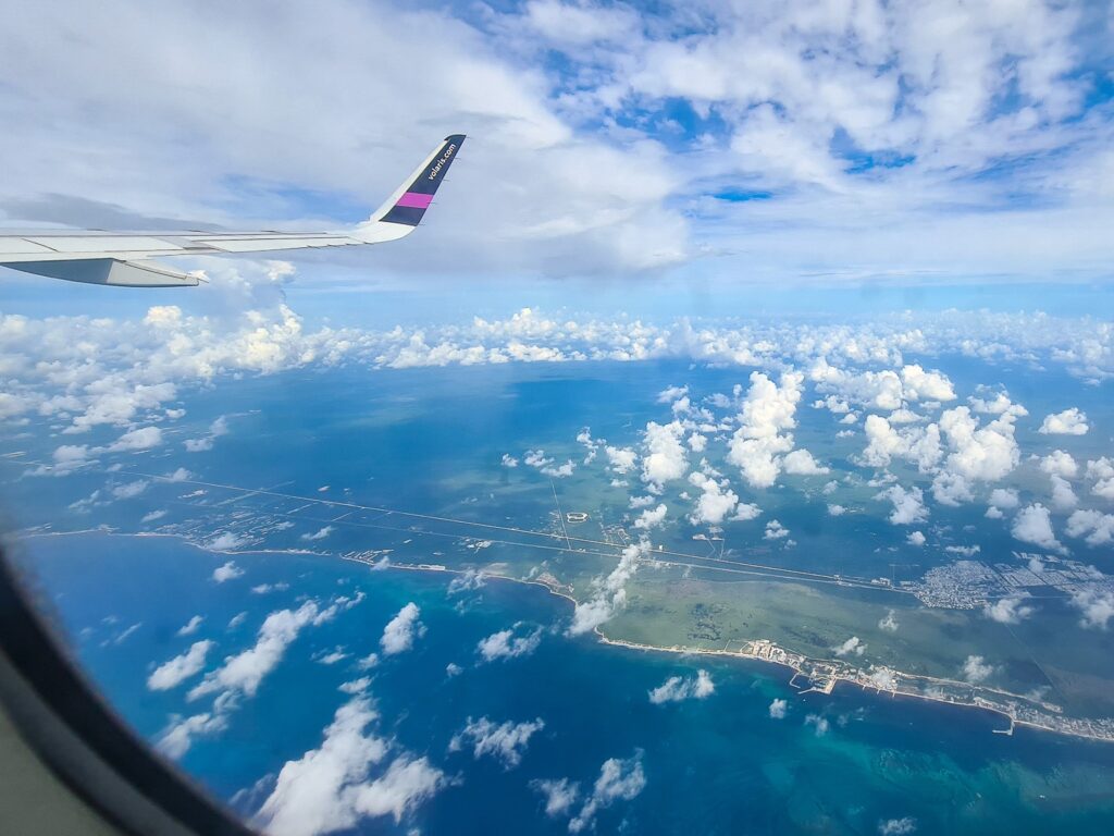 A coastline can clearly be seen out of a plane window. The sky is blue but covered in lots of small white clouds.