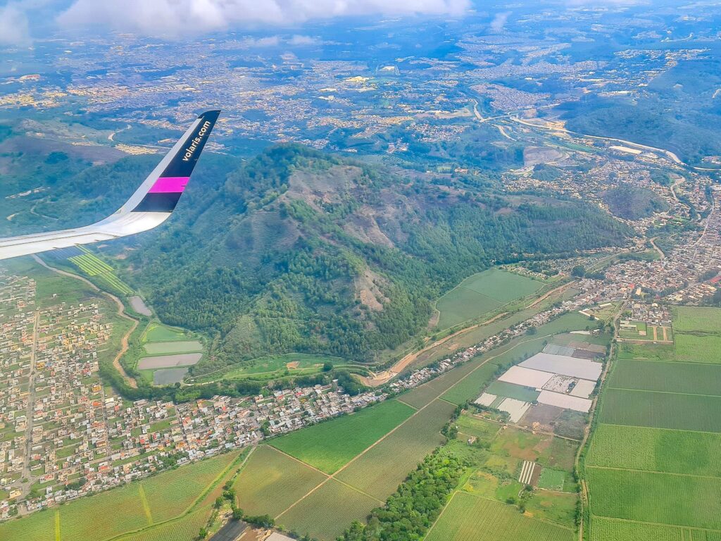 Many trees cover the sides of a tall mountain as we look down at the scene. Surrounding this mountain, many streets of houses and fields can be seen.