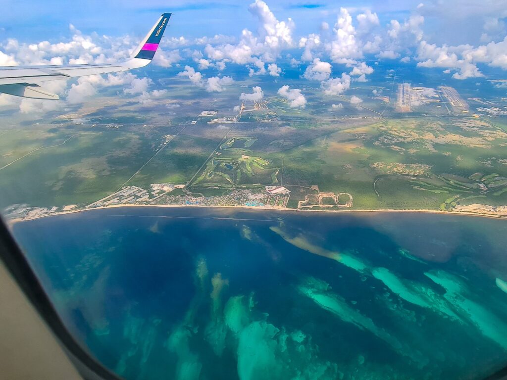 A view out of plane window. We can see a blue-green sea below as it joins with the land. Several small clouds float in the sky.