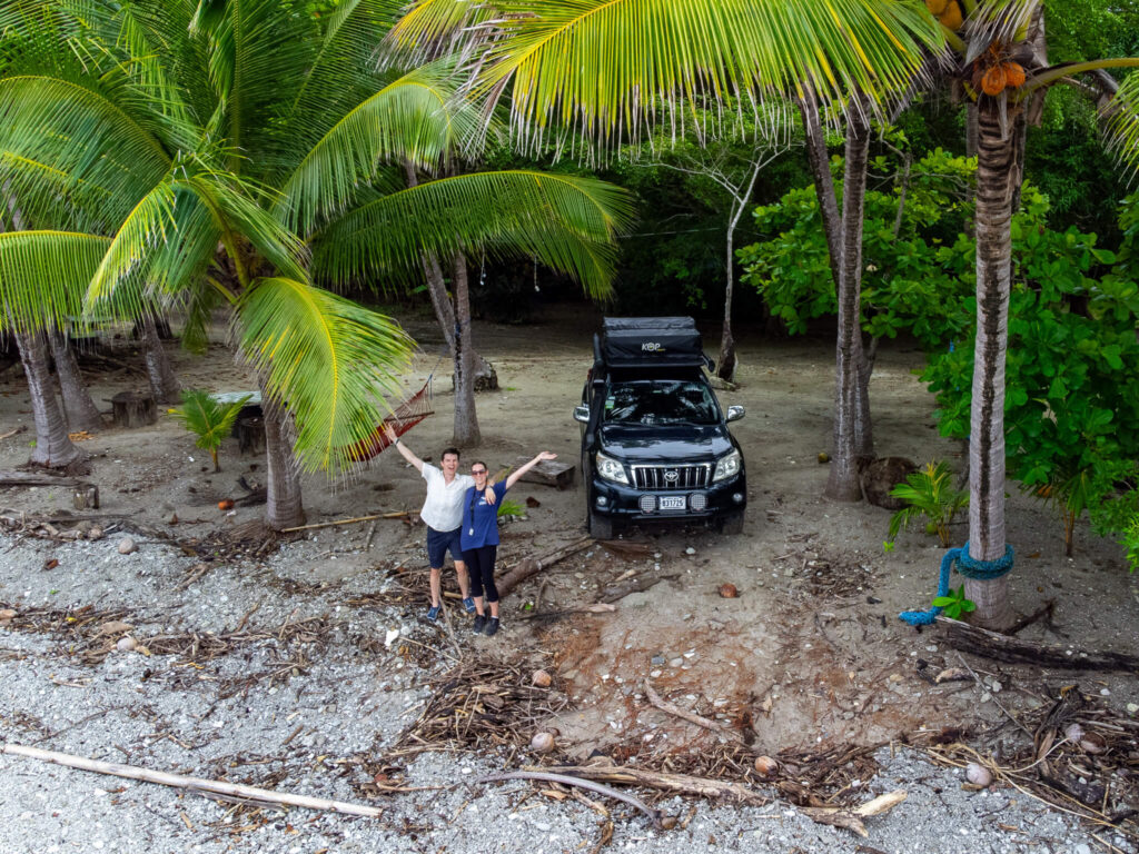 A drone shot of Dan and Lucy standing beside their camper car on a beach with their arms in the air.