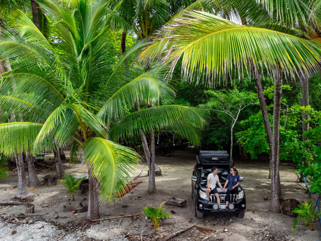 Dan and Lucy holding hands and smiling at each other as they sit on the hood of their 4-wheel-drive. They are parked on a beach under some palm trees.