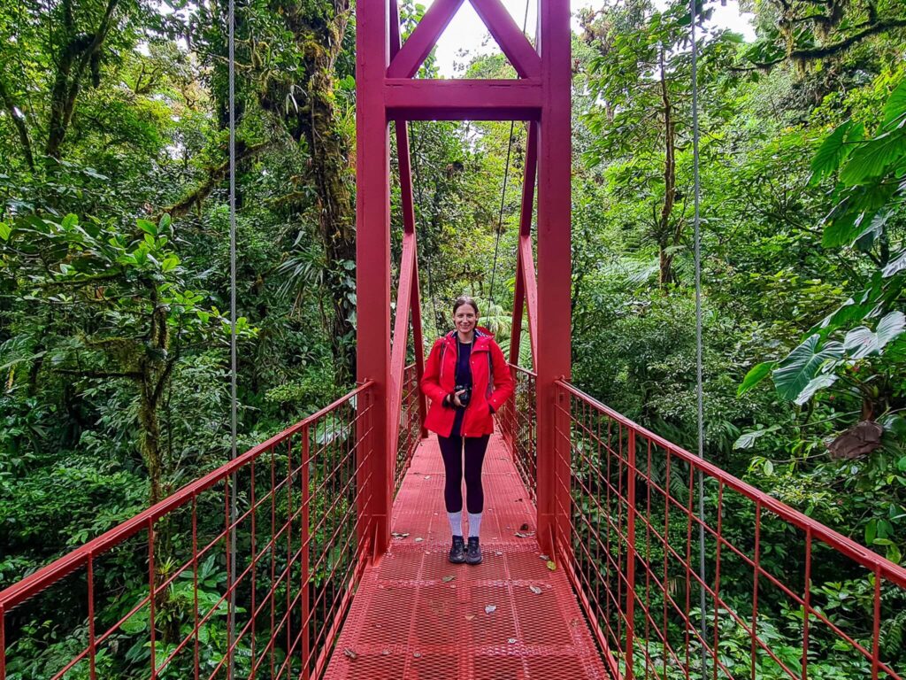 Lucy stands on a bright red metal bridge that stretches across a valley in the Monteverde Cloud Forest.