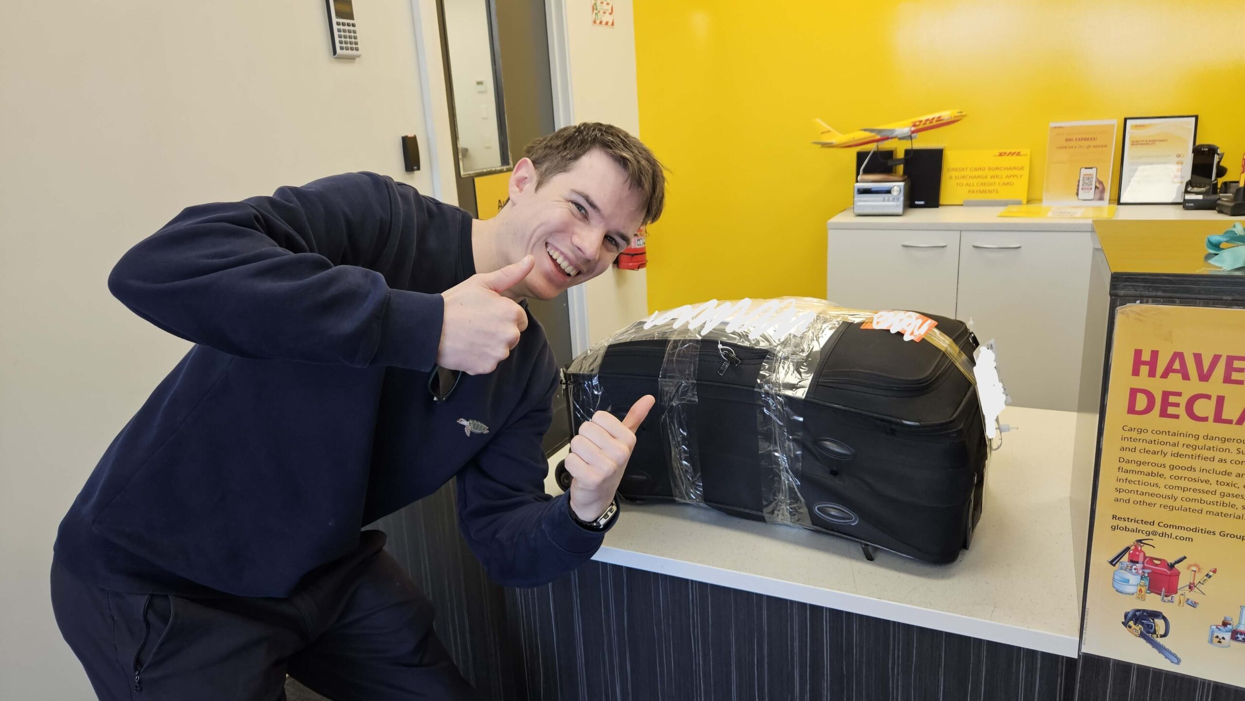 Dan stands next to a tightly wrapped suitcase. He has both thumbs up and a big smile on his face.