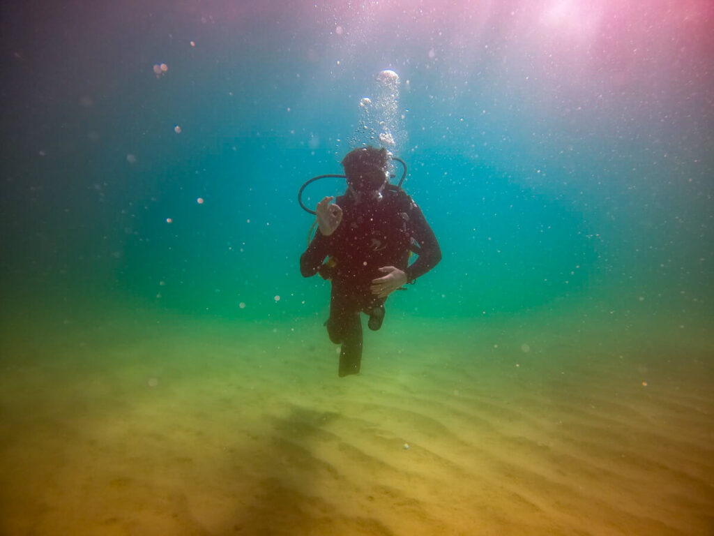 A view underneath the waves: Dan is SCUBA diving and gives the "OK" sign. The water is murky and colourful, spanning the whole spectrum of the rainbow.