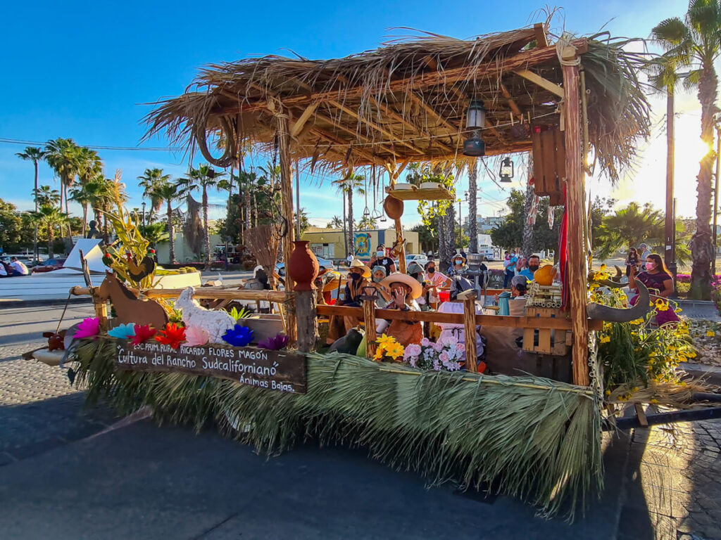 A man waving as he sits on a parade float with his friends. The float is constructed in the style of a traditional Mexican palapa.