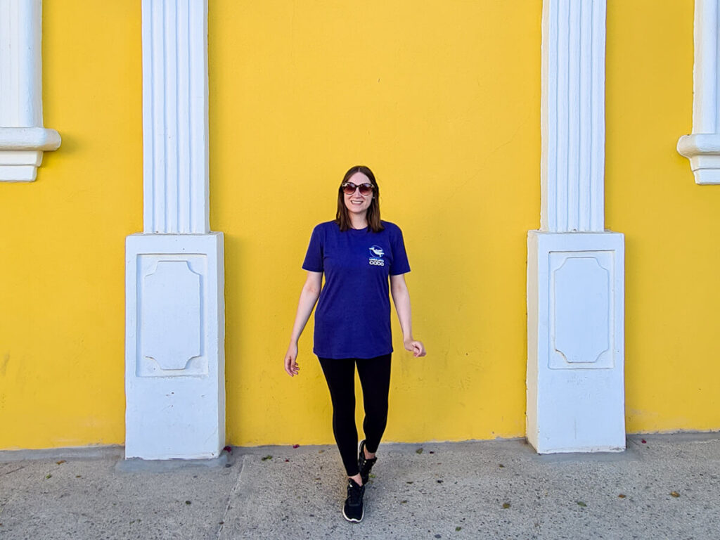 Lucy in front of a bright yellow wall smiling in her sunglasses in the Mexican sunshine.