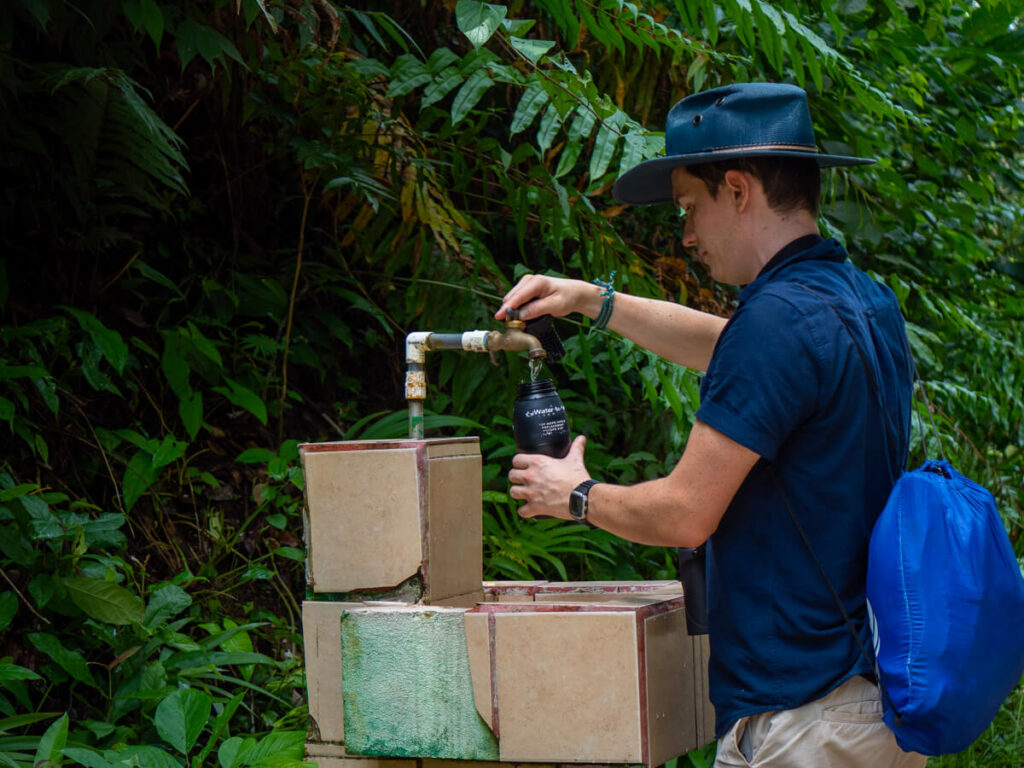 Dan standing at an outdoor tap, refilling his reusable Water-to-Go bottle. He is wearing a large blue hat and is surrounded by greenery.