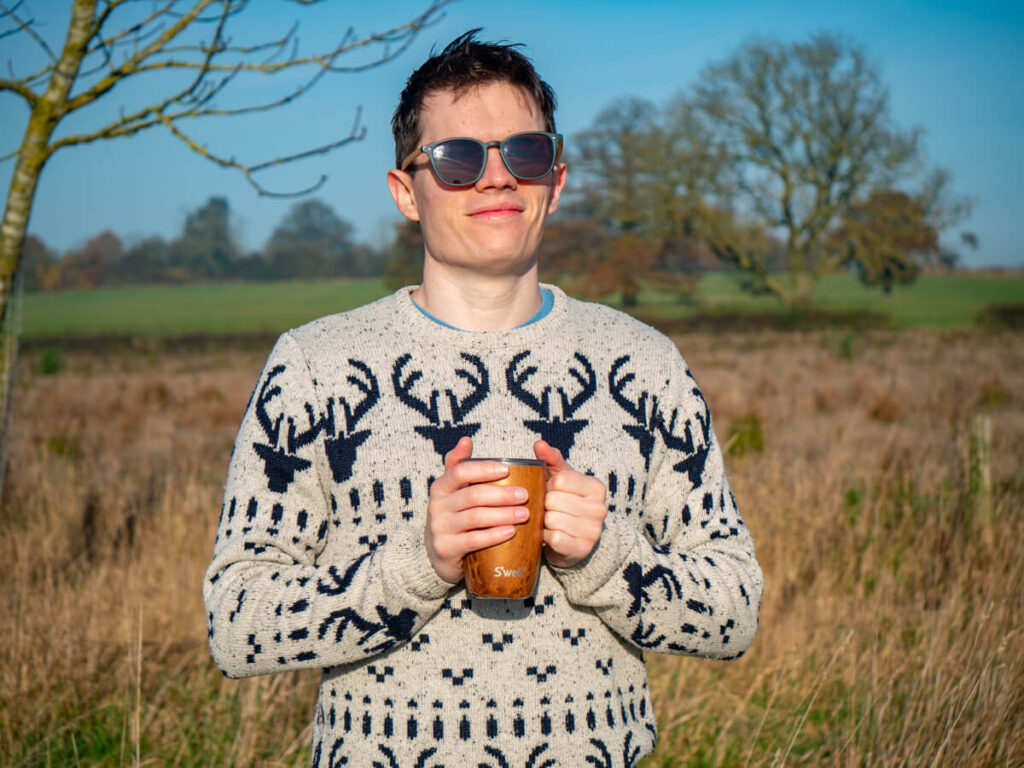 Dan smiling in the sunshine holding a coffee cup with a wooden design. He is wearing sunglasses and a black-and-white Christmas jumper.