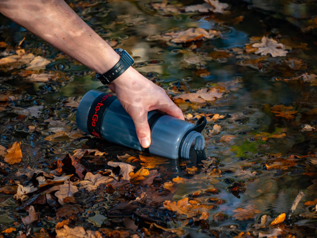 Filling up a translucent water bottle from a stream covered with leaves. The words "Water-to-Go' are printed towards the bottom.