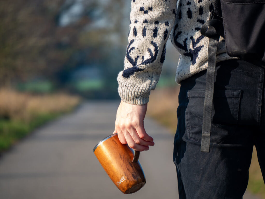 Dan holding a reusable coffee cup loosely by its handle. Straight ahead is a path through the countryside.