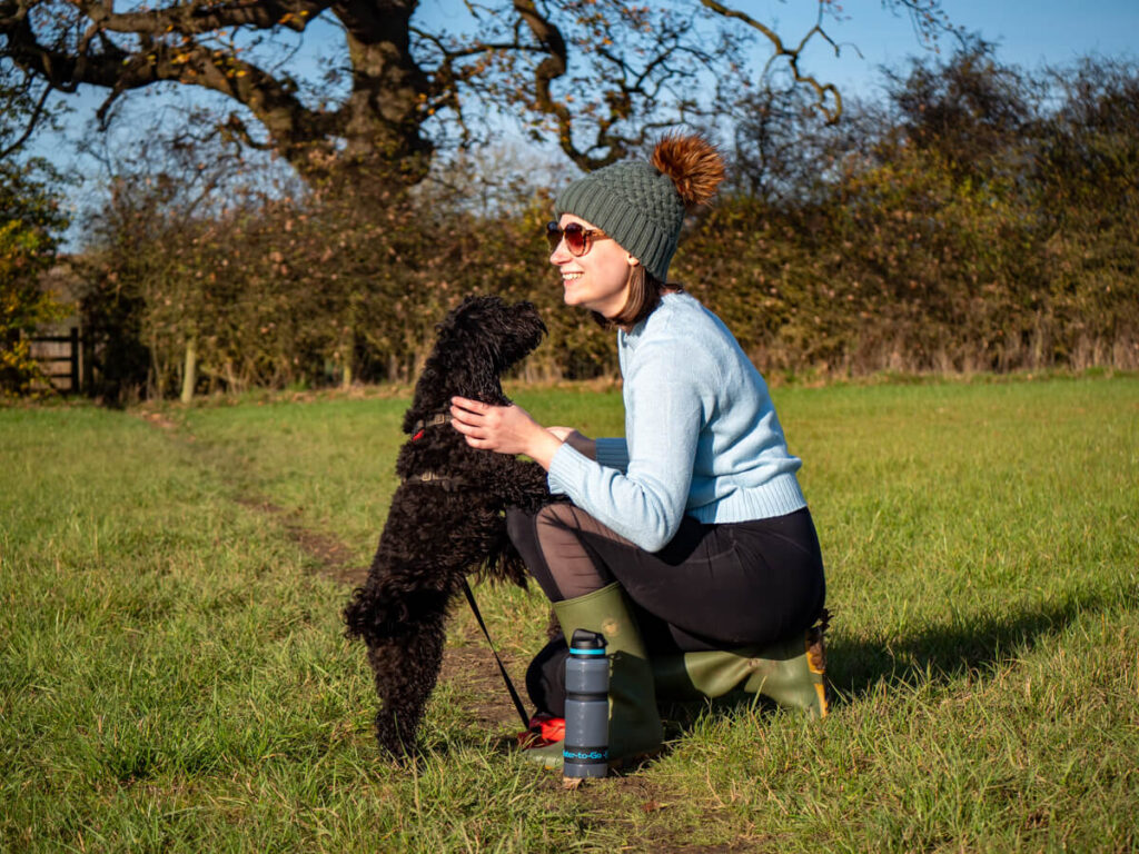 A dog standing on its hind legs greeting Lucy as she bends down smiling in the sunshine. A water bottle is resting in front of Lucy's wellington boots.