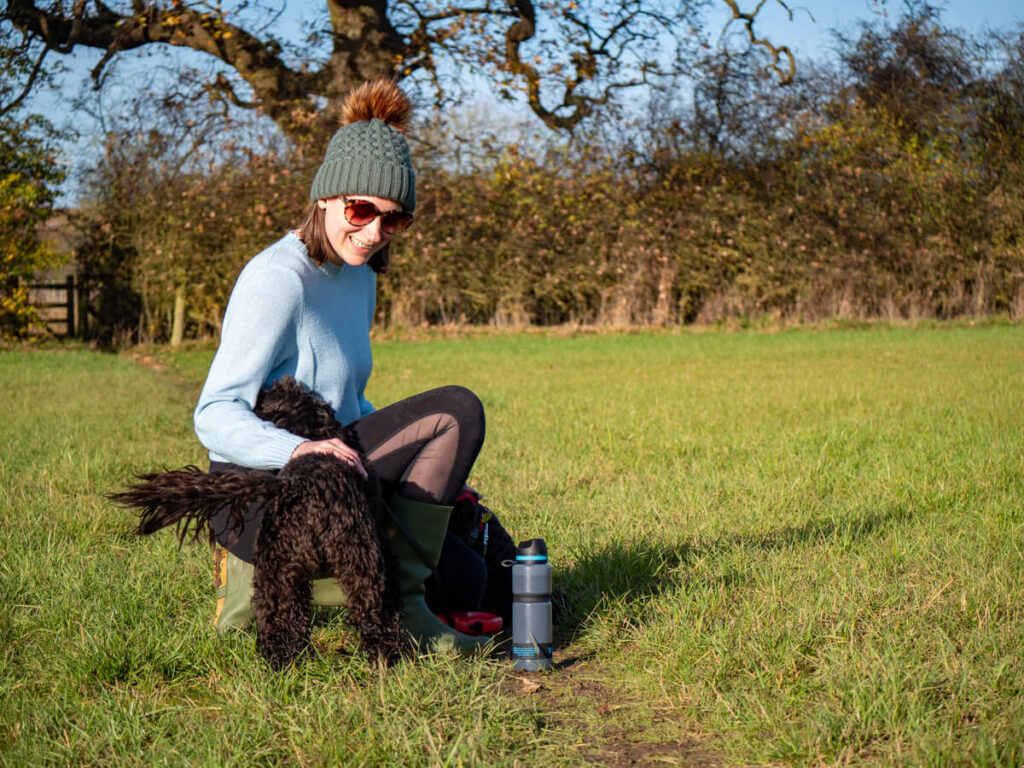 Lucy smiling as she pets a fluffy dog in a field. She is wearing a bobble hat and is knelt beside a blue Water-to-Go bottle.