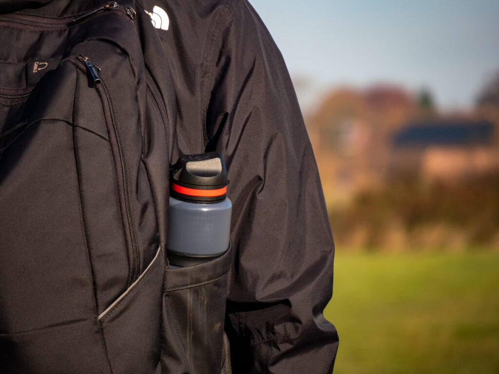 A filter water bottle with a red design donned in the side-pocket of a backpack.