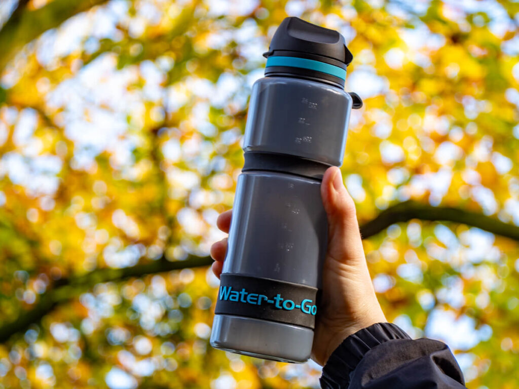 A translucent, reusable water bottle held up in front of a sunny forest scene.