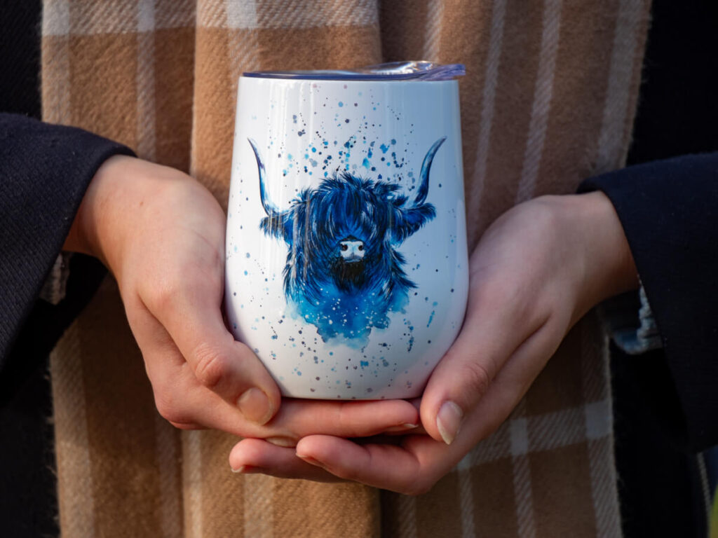 Lucy holding a reusable coffee cup that has a blue highland cow design painted on it.