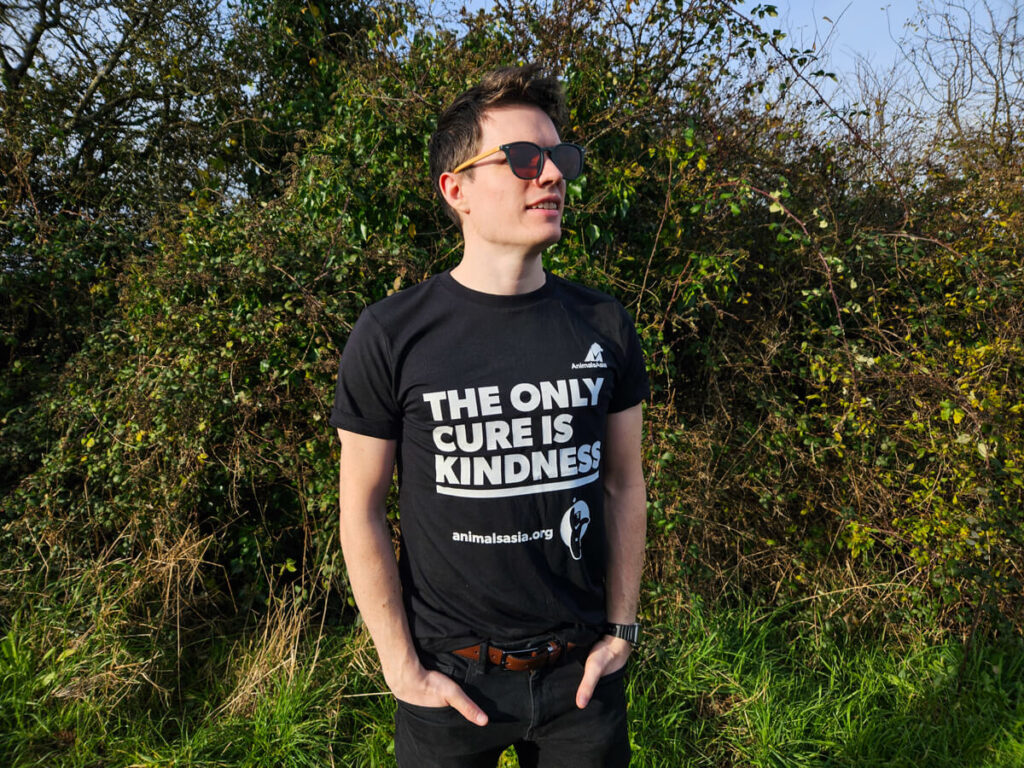 Dan stands in front of some dark green bushes. He is wearing sunglasses and a black t-shirt with the words "The only cure is kindness. animalsasia.org" printed on the front.