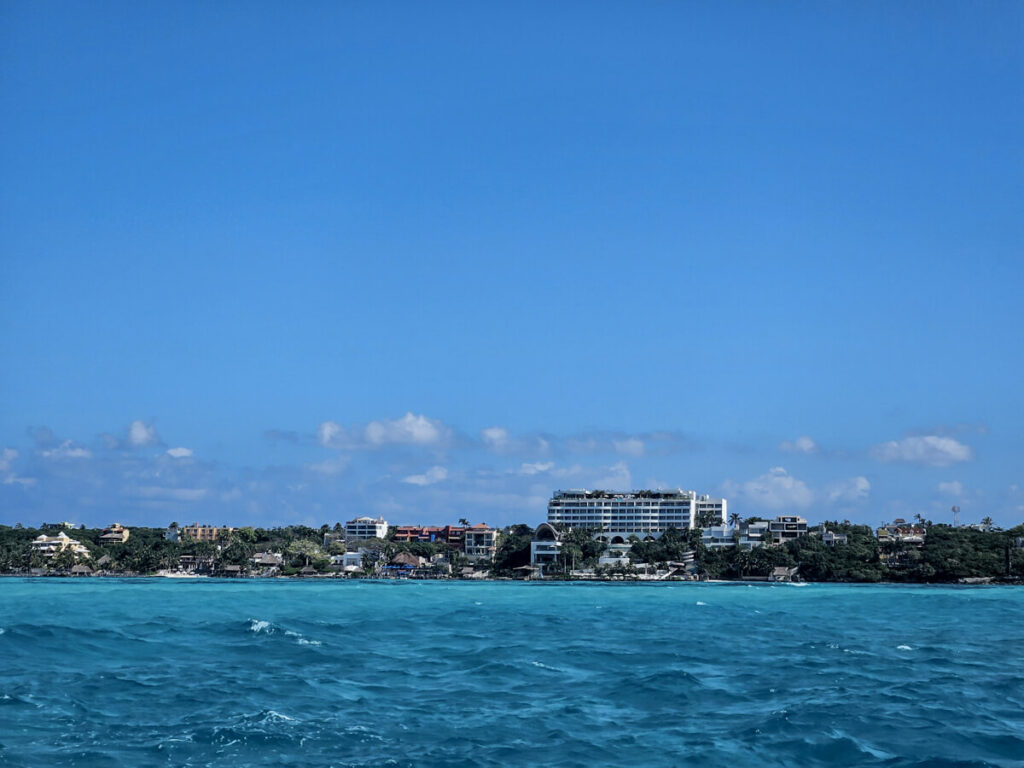 The coast of Cancun as viewed from a boat. There are many large buildings with trees and other greenery between them.