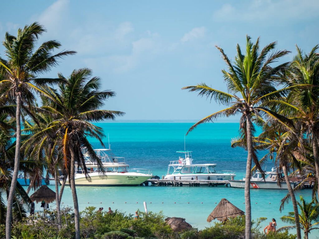 Boats docked in the light blue waters of Isla Contoy. Palm trees line either side of the view and a view tourists can be seen on the beach nearby.